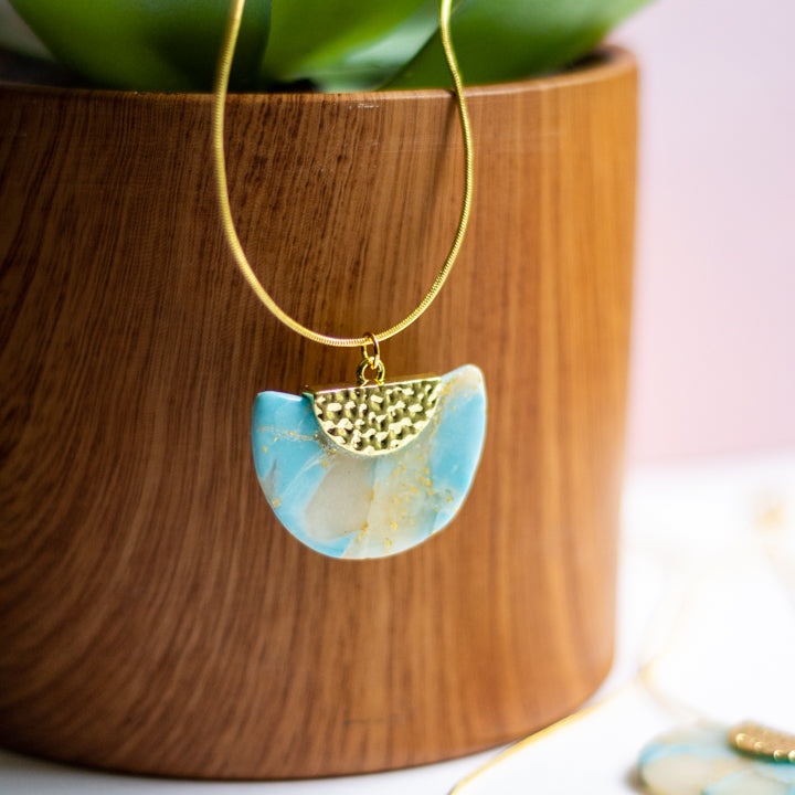 Necklace with a blue and gold pendant in front of a wooden planter with green leaves.