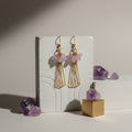 Gold earrings with pink gemstones on a white stand with amethyst crystals.
