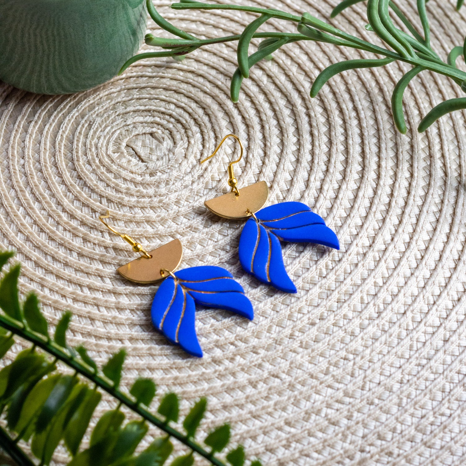 Handmade blue statement earrings with gold-plated hooks and layered polymer clay leaf shapes, lightweight geometric drop earrings styled on a woven natural background.