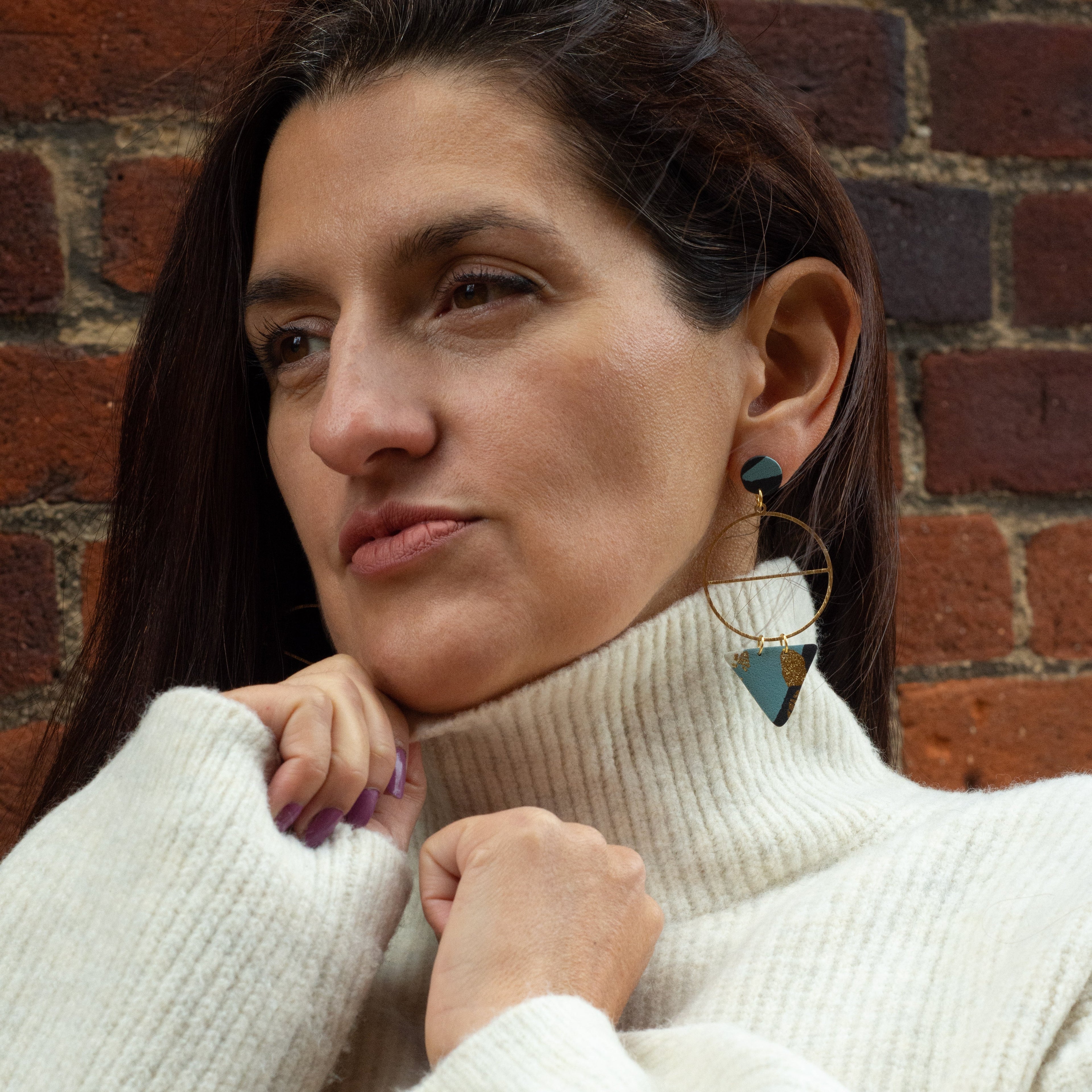 Woman wearing a white turtleneck sweater against a brick wall and wearing blue and black earrings