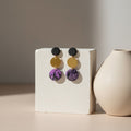 Pair of earrings with purple and gold components on a white stand, with a beige vase in the background.