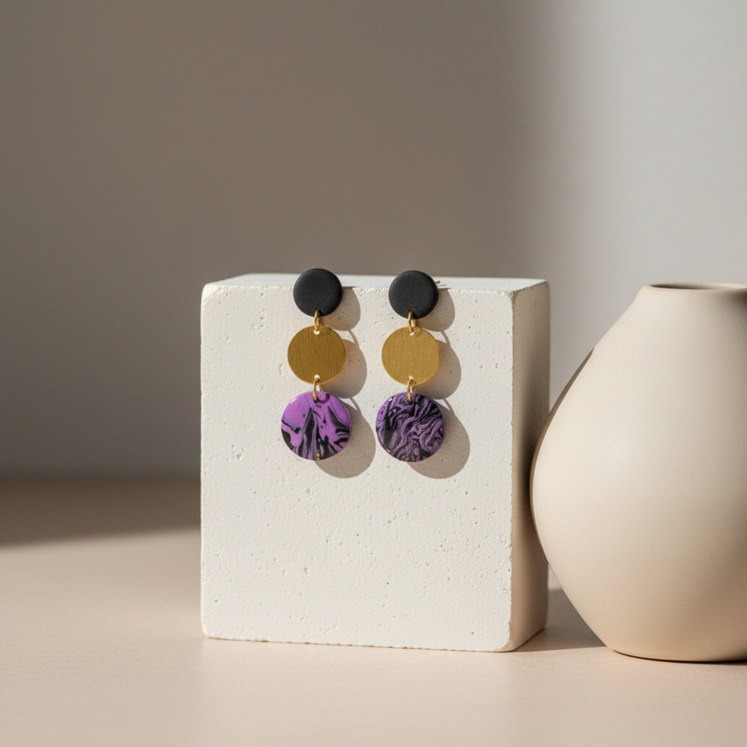Pair of earrings with purple and gold components on a white stand, with a beige vase in the background.