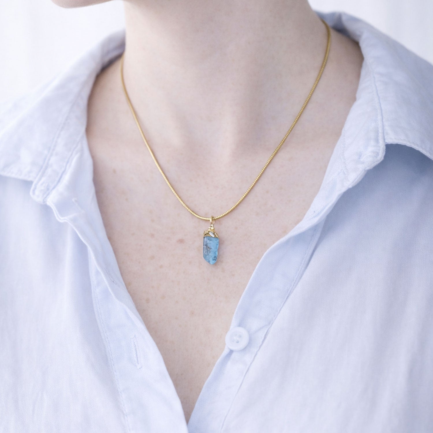 Gold necklace with a blue apatite pendant worn by a person in a white shirt.