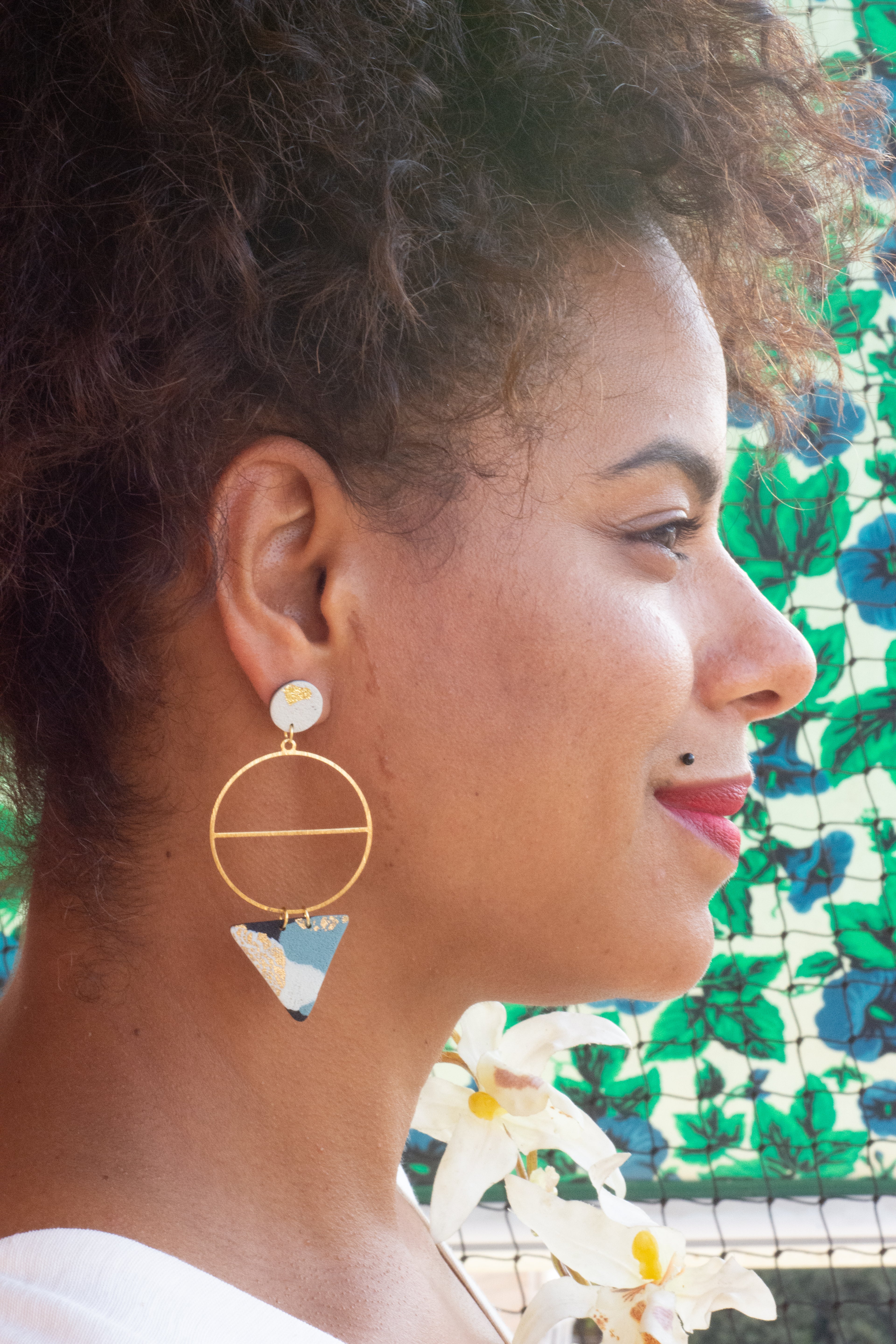 Woman wearing gold hoop earrings with a geometric design, against a colorful floral background.