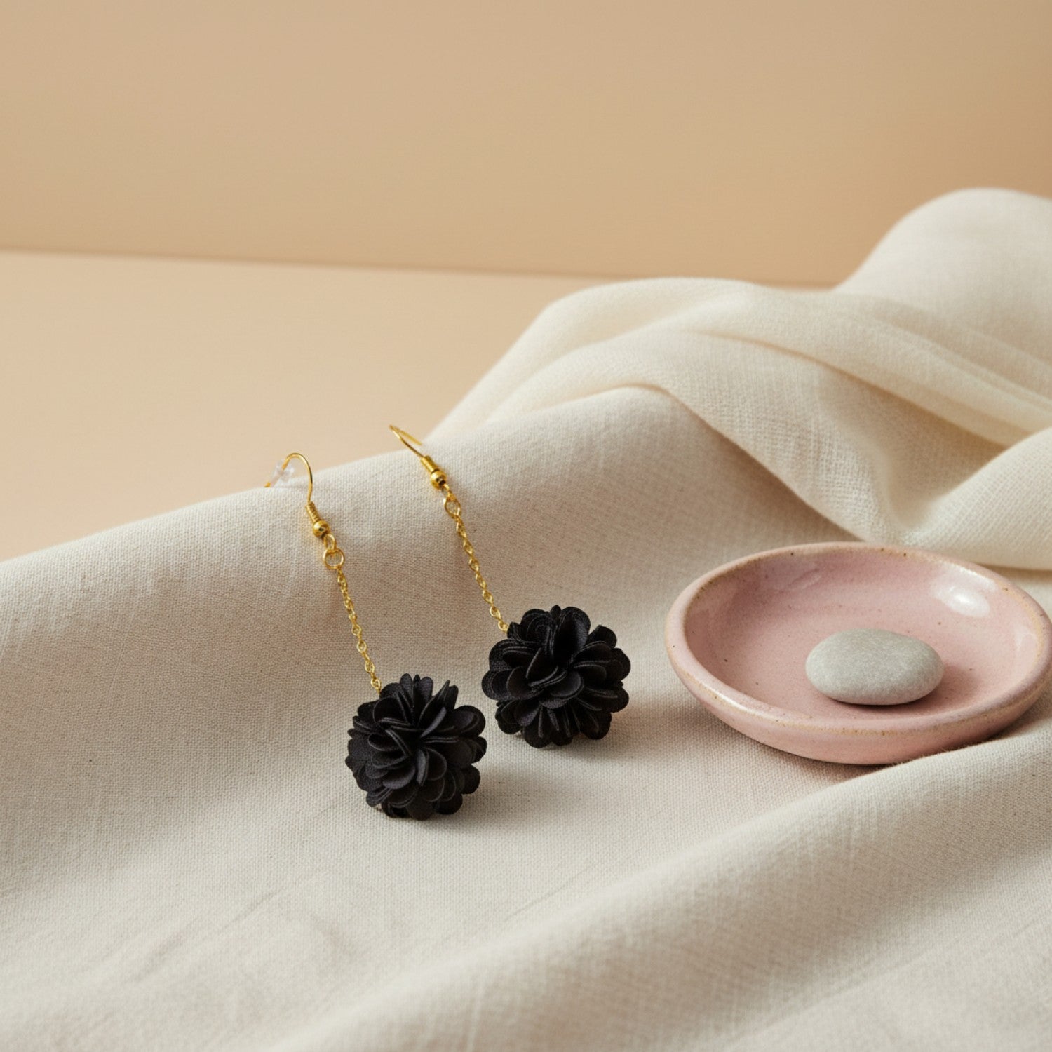 Black floral earrings with gold chains on a beige surface with a pink dish.