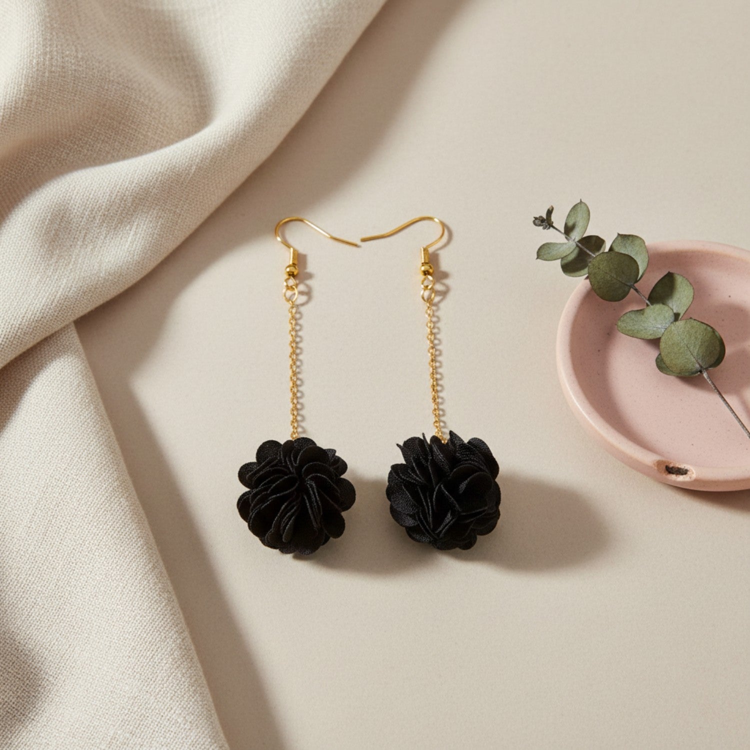 Black flower-shaped earrings with gold chains on a beige surface with a pink dish and green leaves.