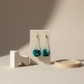 Teal pompom earrings on a white block with a beige background