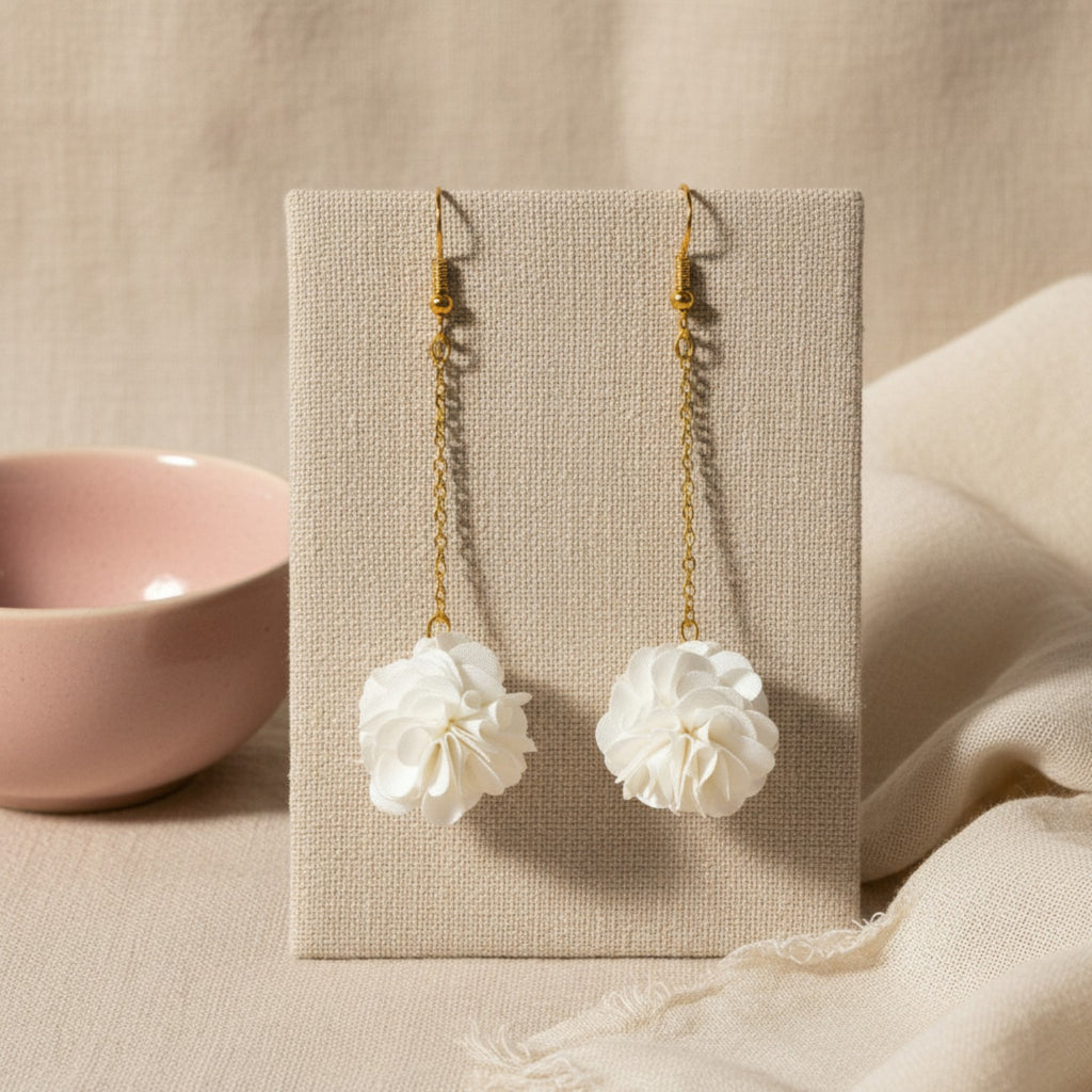 Pair of white pompom earrings on a beige card with a pink bowl in the background