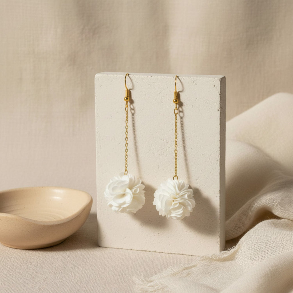 Pair of white pompom earrings on a beige card with a pink bowl in the background