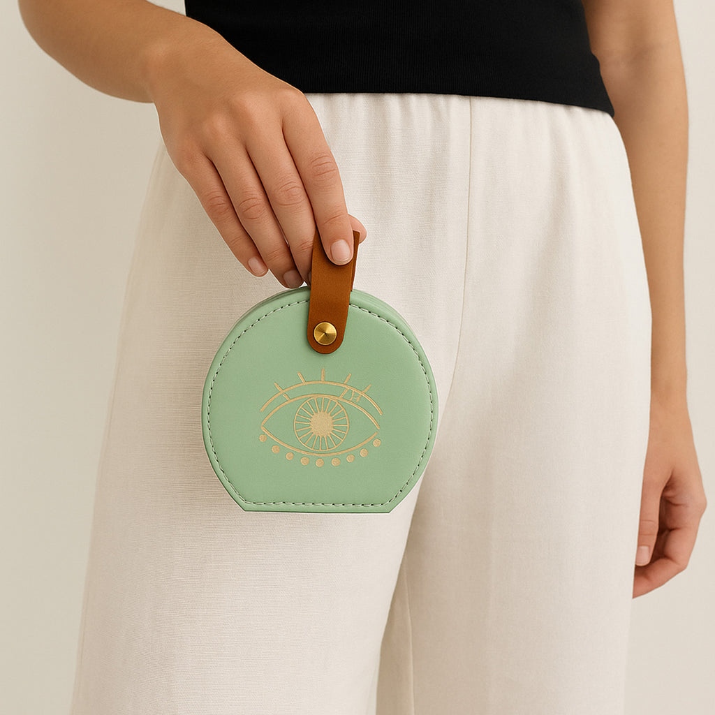 Green round mini jewellery box with eye design held by a woman with white troussers. 