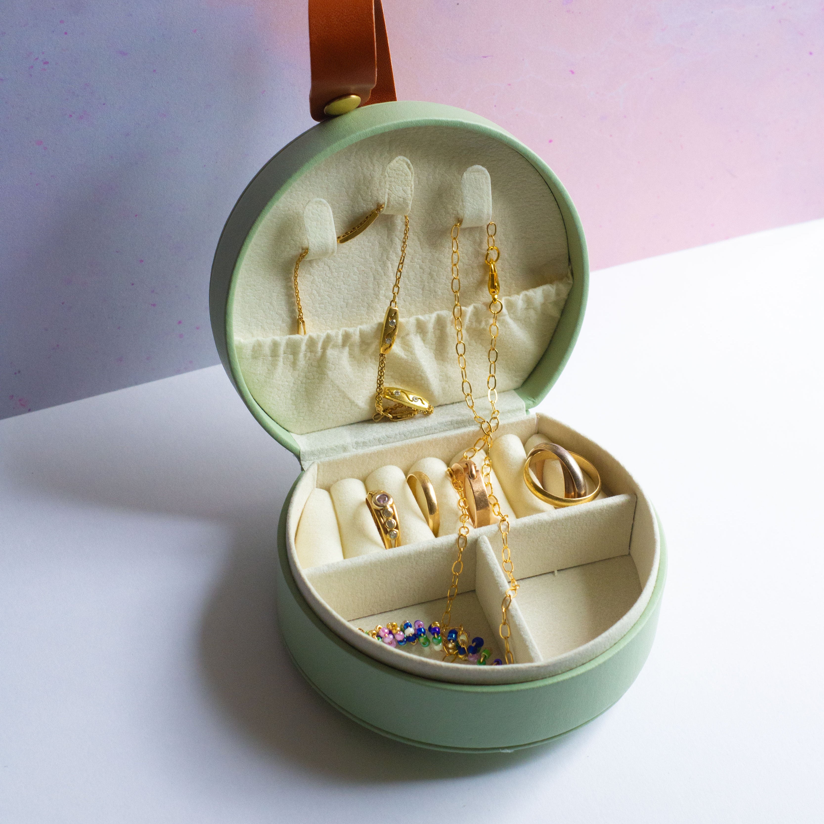Open green round mini jewellery box with rings and necklaces on a pink background