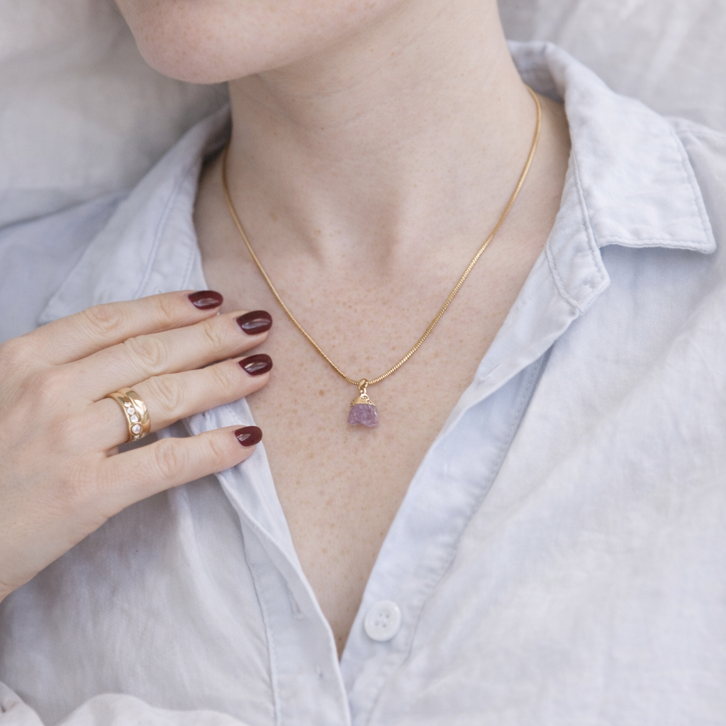 Woman wearing a gold necklace with a rose quartz pendant, holding her hand to her chest.