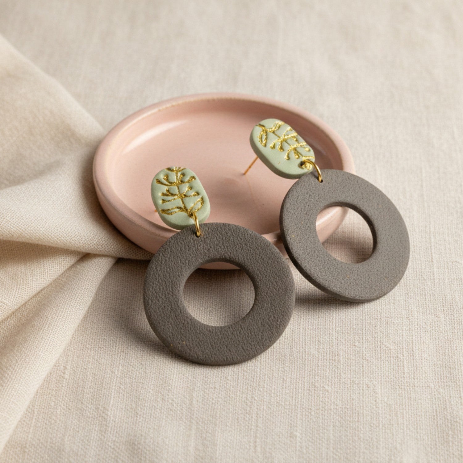 Pair of earrings with gray circular pendants and green leaves on a beige surface