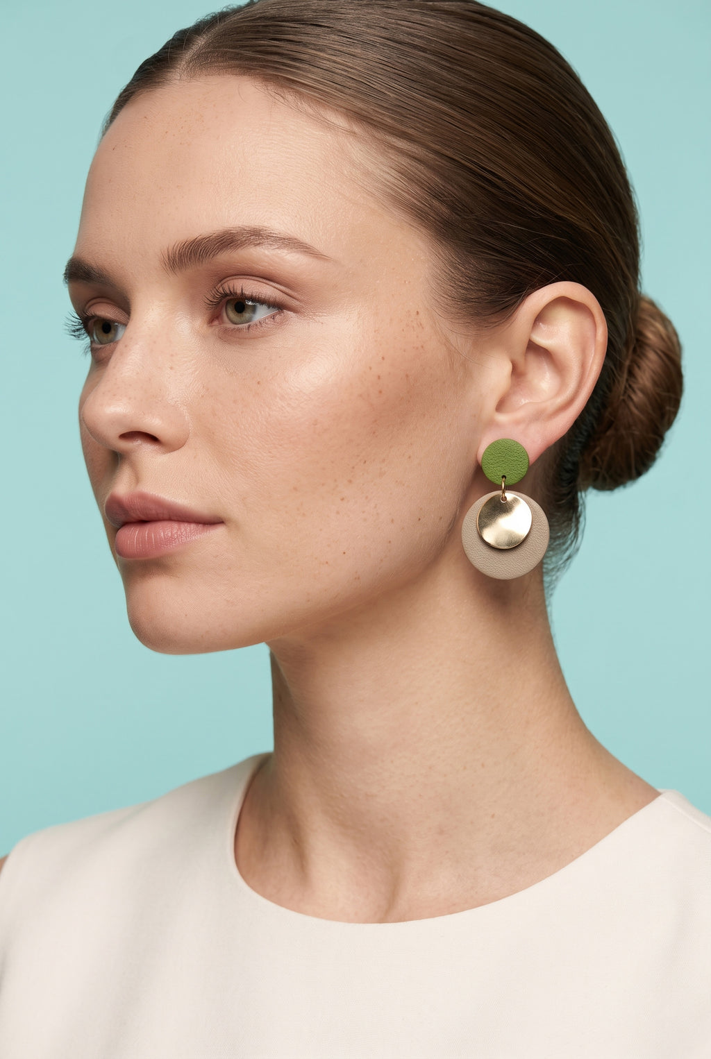A close-up side profile of a woman wearing a pair of statement dangle earrings. The earrings feature a small olive green disc on top of a larger, matte metallic gold disc.