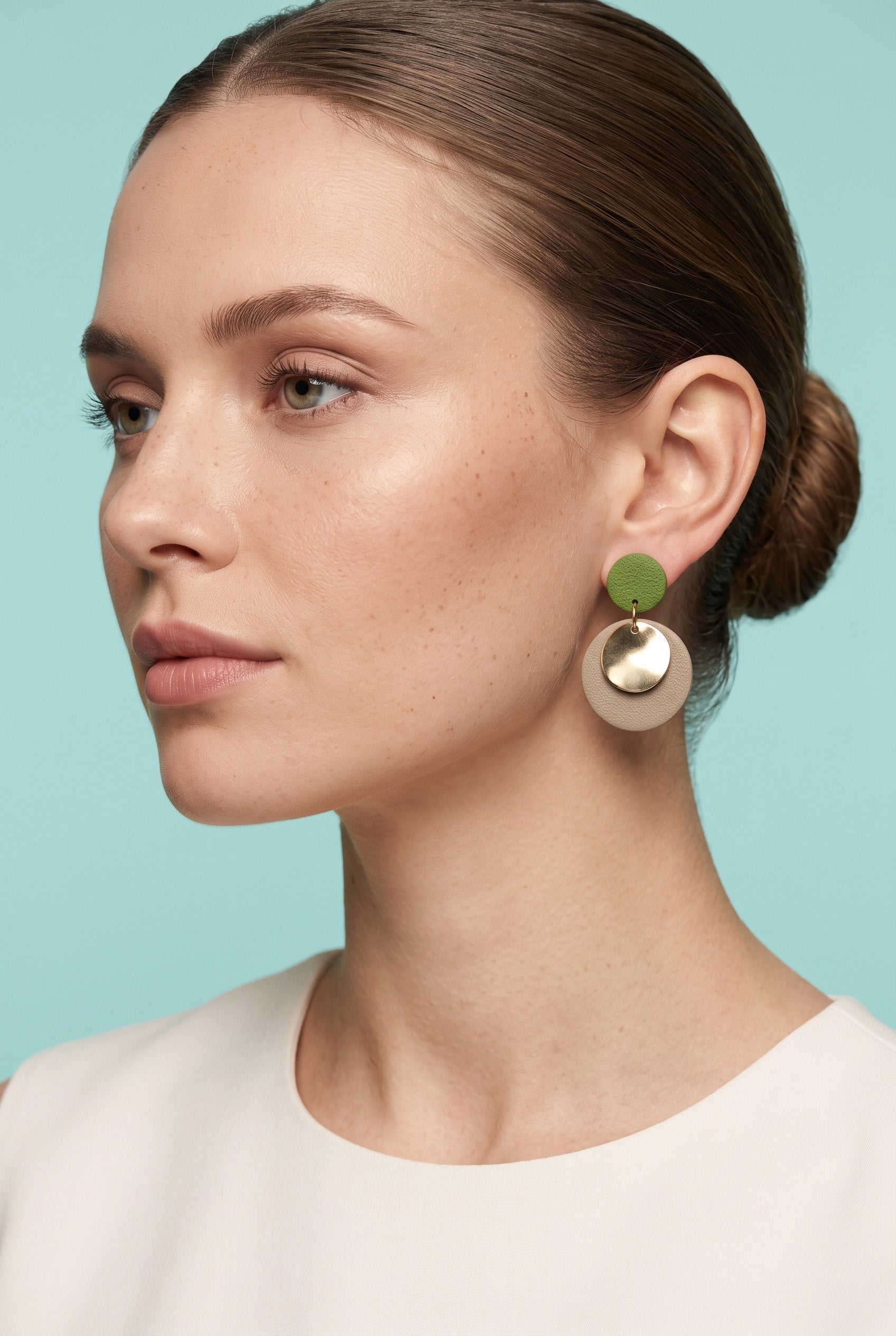 A close-up side profile of a woman wearing a pair of statement dangle earrings. The earrings feature a small olive green disc on top of a larger, matte metallic gold disc.