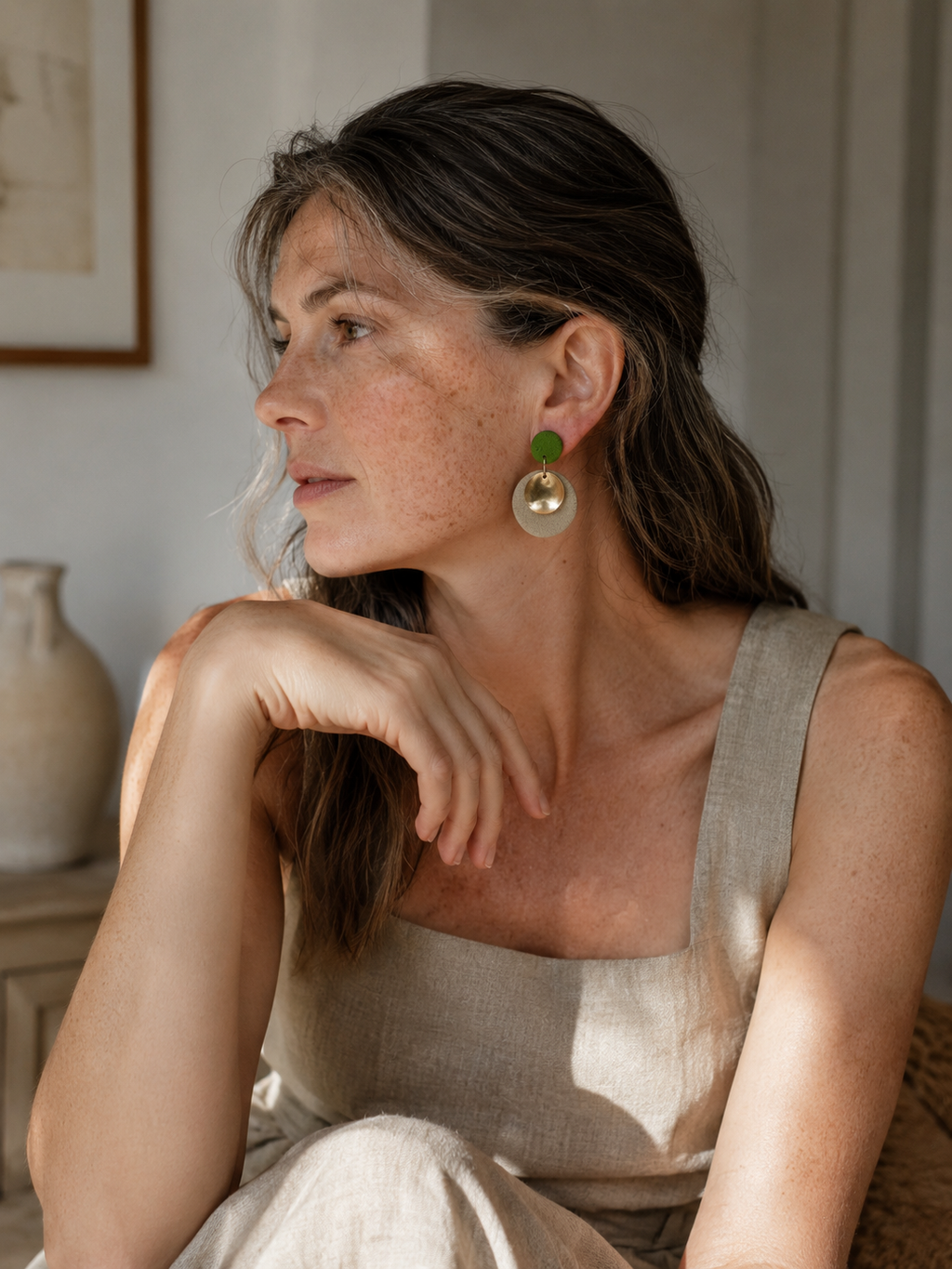 A woman in a beige linen top wearing a pair of large, bohemian-style earrings with green and cream elements, posed in a softly lit indoor setting.
