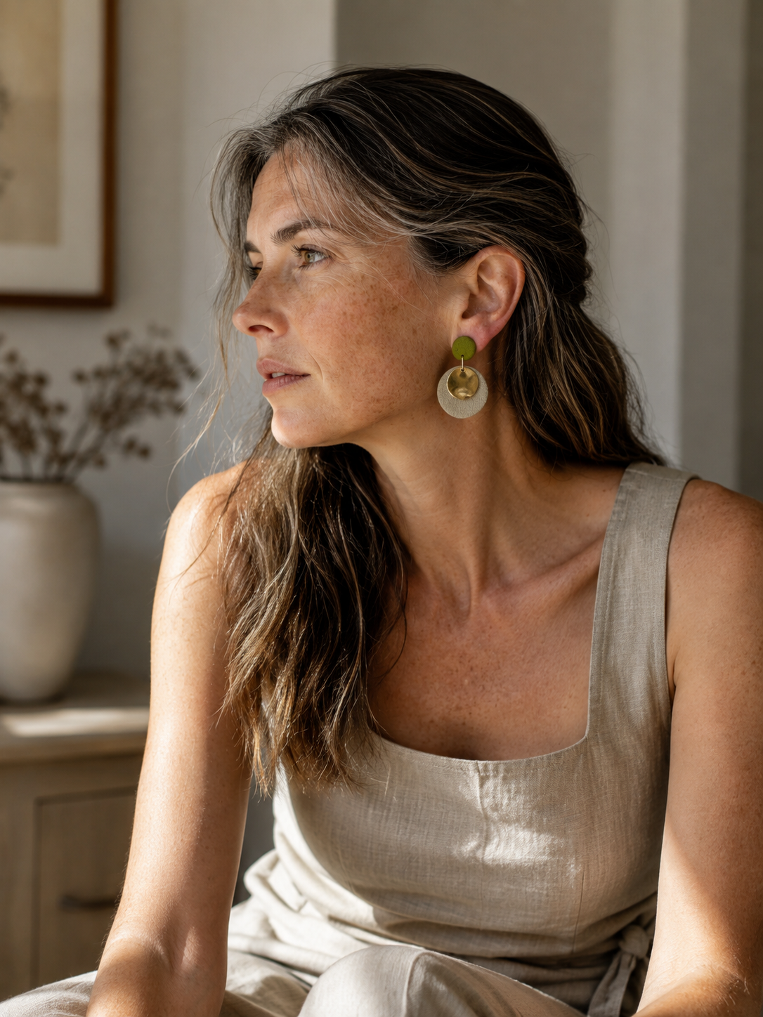 A woman with long brown hair wearing a beige linen top and matching bottoms, looking to the side and wearing a large circular gold and green earring.