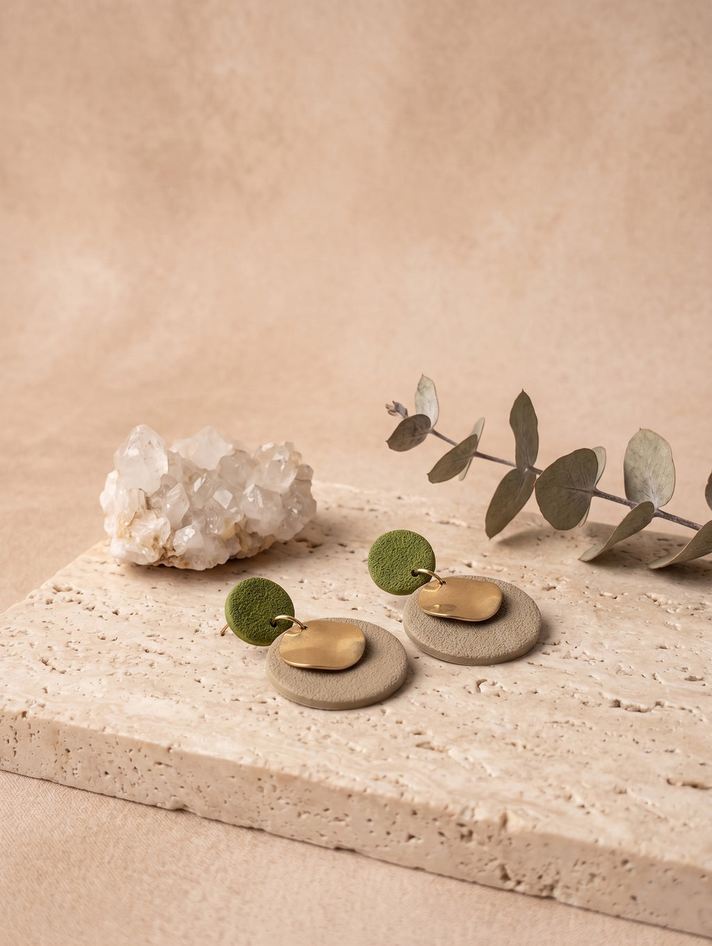 Two minimalist seed-shaped earrings with olive green and brass gold elements, displayed on a textured stone slab with a crystal cluster and eucalyptus branch against a warm beige background.