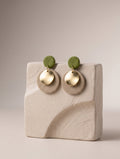 A pair of handmade dangle earrings displayed on a textured beige ceramic block. The earrings feature olive green circular studs, gold-toned disc charms, and beige textured circular drops.