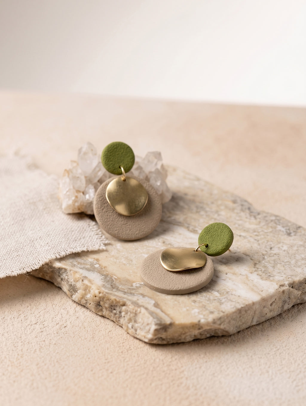 Two minimalist dangle earrings displayed on a textured stone slab. Each earring features a round beige textured disc, a small polished gold leaf-shaped charm, and a small round sage green disc at the top.