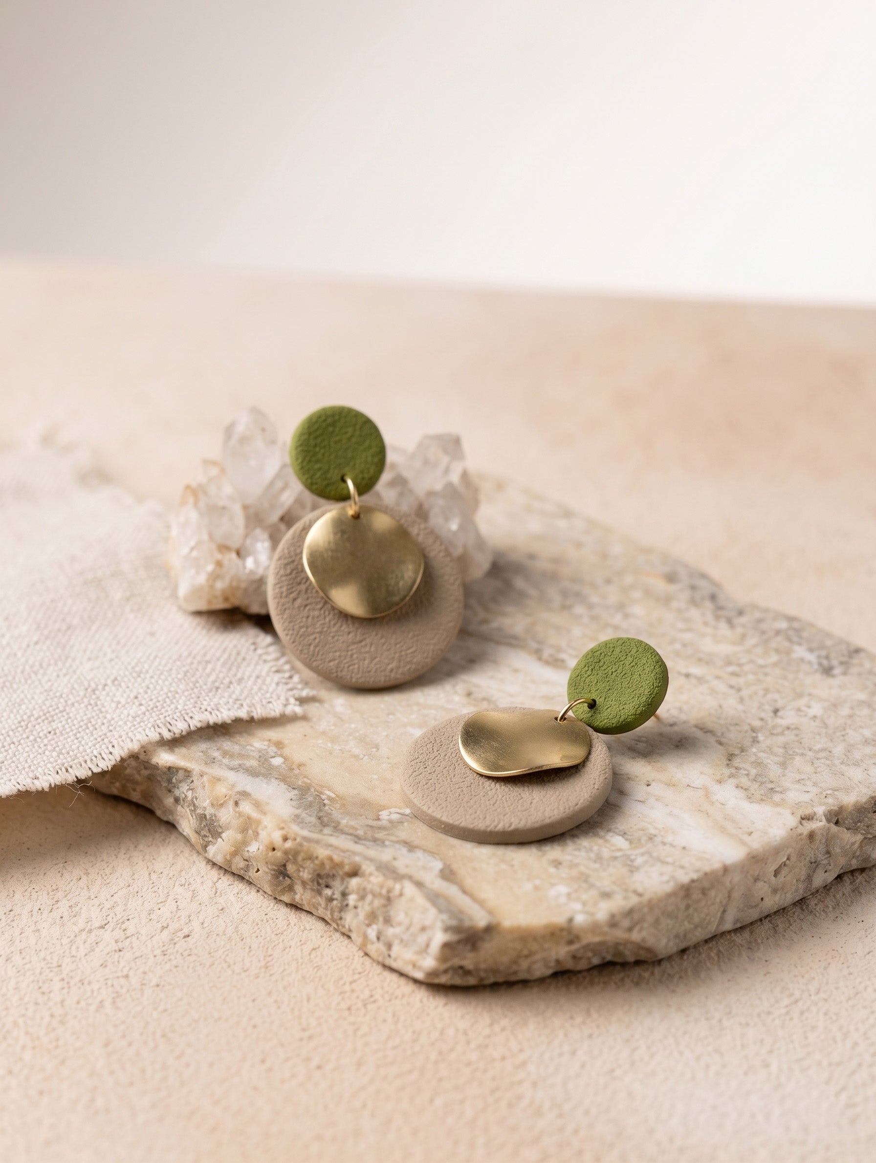 Two minimalist dangle earrings displayed on a textured stone slab. Each earring features a round beige textured disc, a small polished gold leaf-shaped charm, and a small round sage green disc at the top.