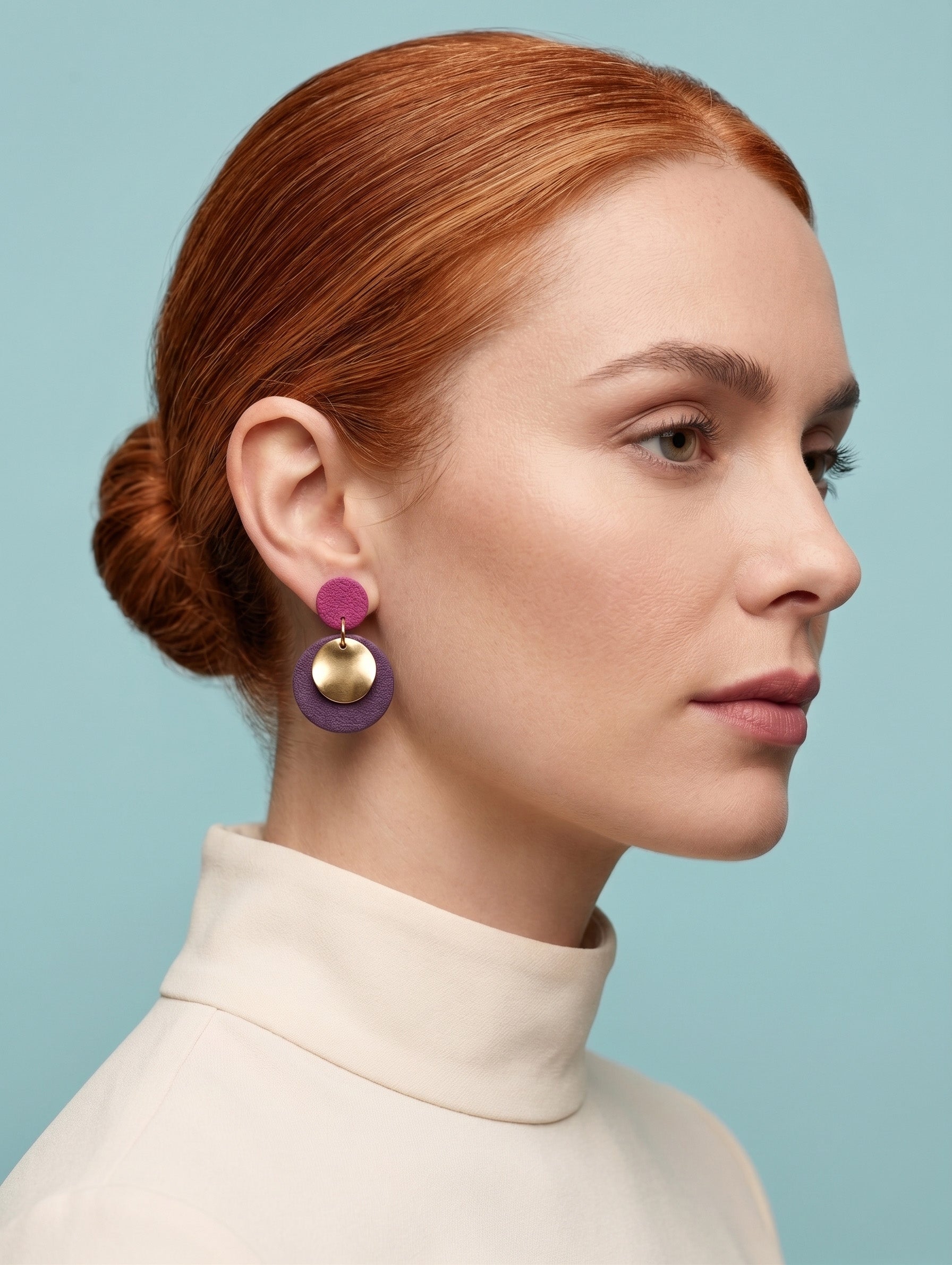 A close-up side profile of a woman with red hair in a bun, wearing a pair of colorful geometric drop earrings with purple, gold, and magenta discs, set against a solid light blue background.