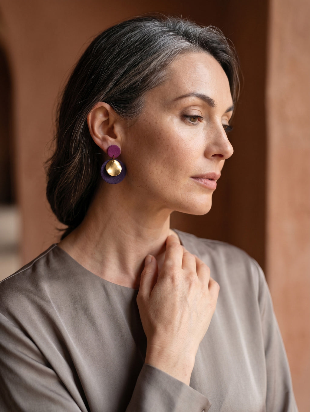 A woman in profile wearing a pair of distinctive multi-colored drop earrings with purple, gold, and dark purple elements, set against a warm, blurred terracotta background.