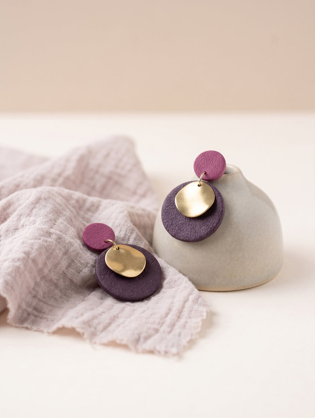 A pair of handmade disc earrings with purple and gold metal layers, displayed on a light beige ceramic stand and a textured pale pink cloth against a warm cream background.