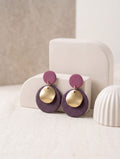 A pair of handmade dangle earrings with textured purple circular discs and shiny gold disc charms, displayed on a cream-colored geometric stand.