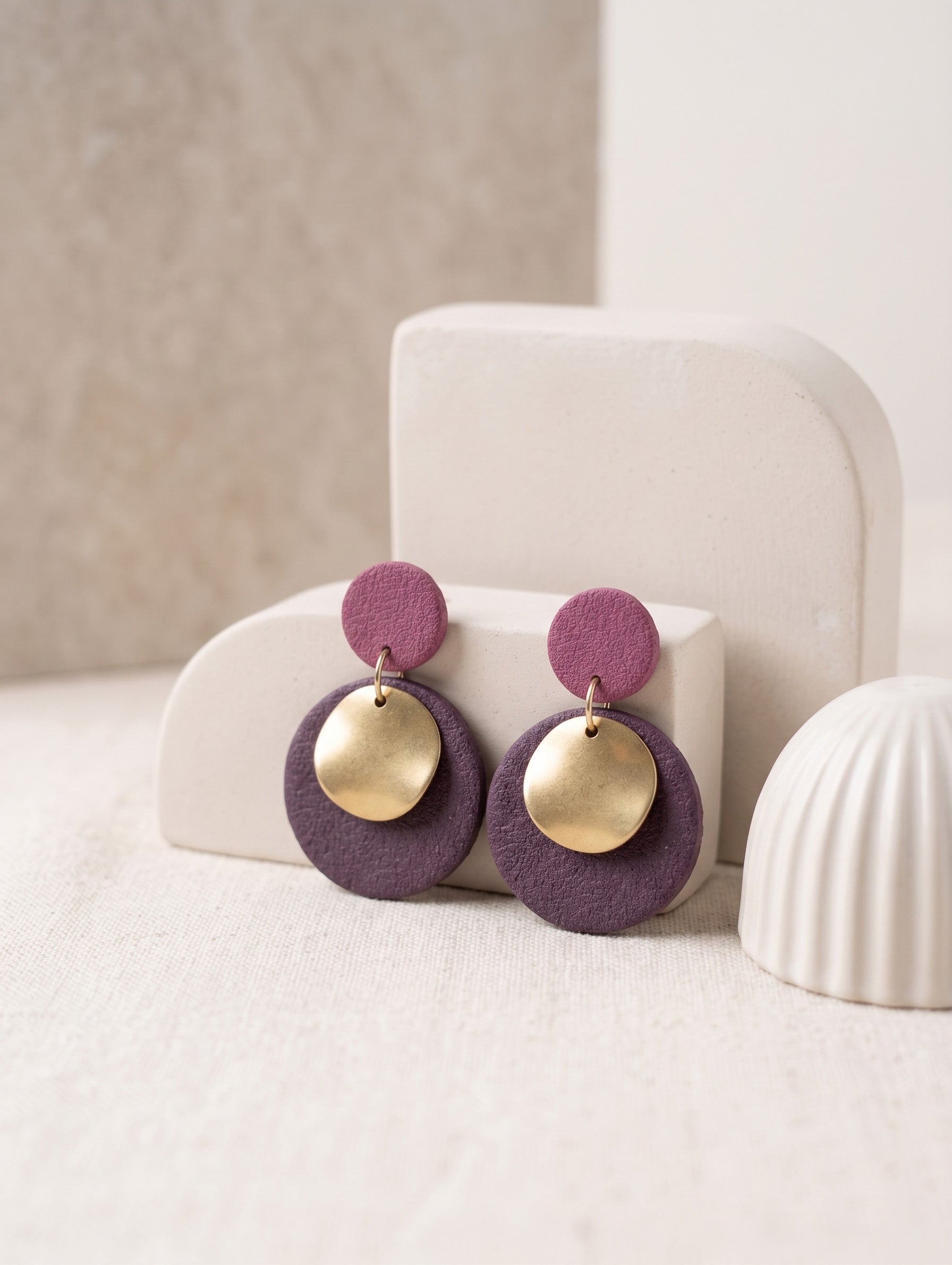 A pair of handmade dangle earrings with textured purple circular discs and shiny gold disc charms, displayed on a cream-colored geometric stand.