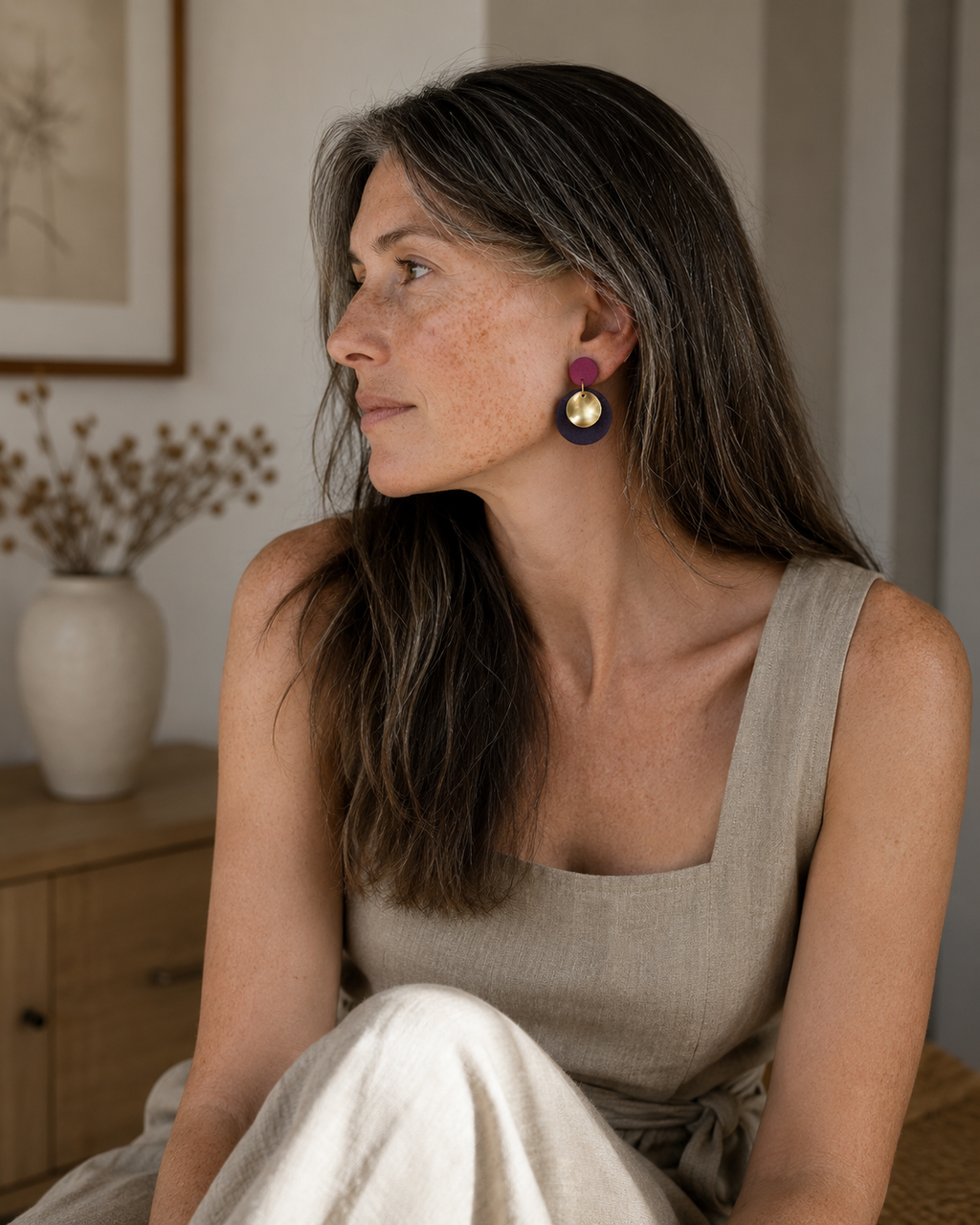 A woman with long brown hair wearing a beige linen top and light-colored pants, looking to the side while wearing a pair of colorful geometric drop earrings with purple, gold, and navy blue.