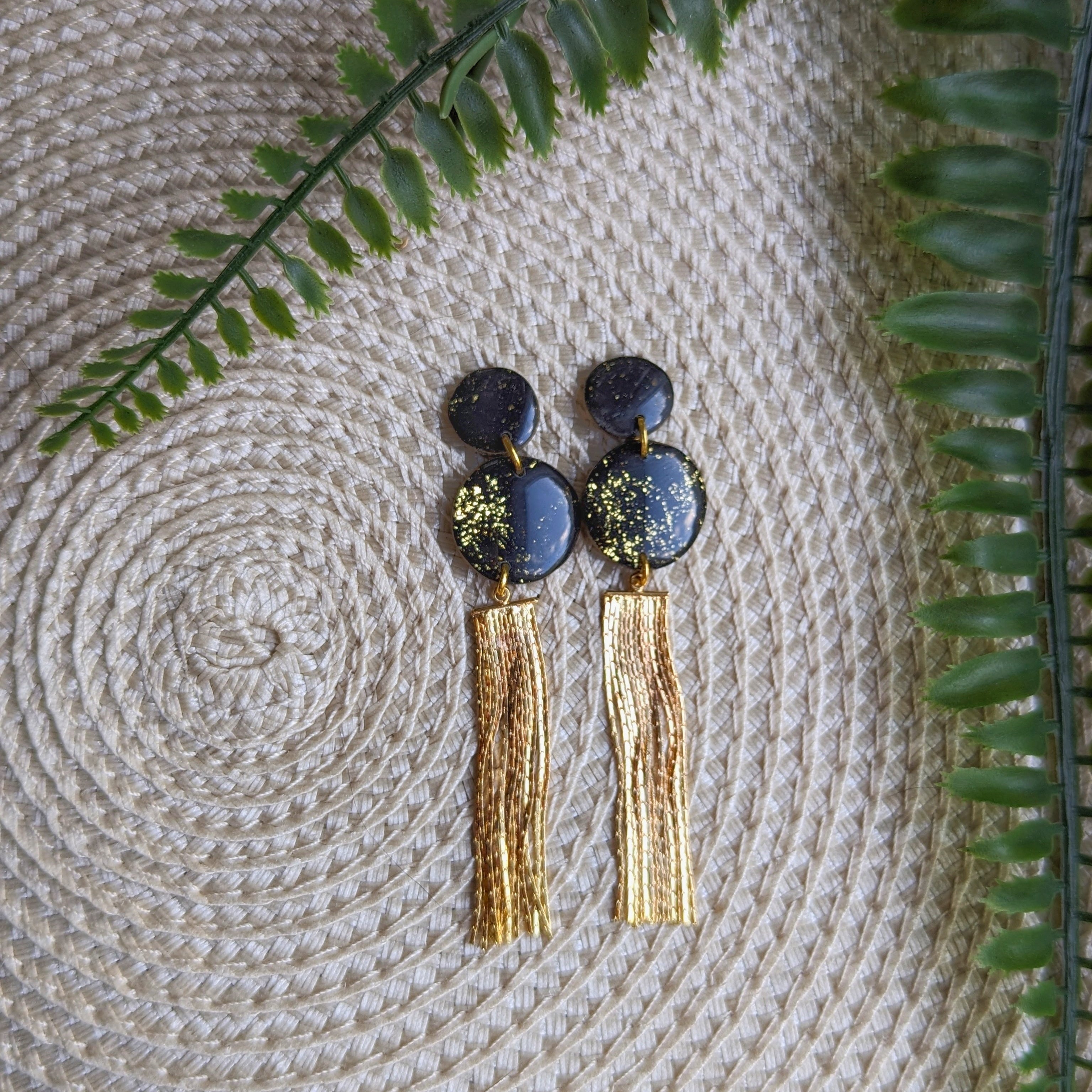 Handmade black and gold statement tassel earrings with gold-plated chain fringe and speckled polymer clay discs, elegant lightweight drop earrings on a white background.