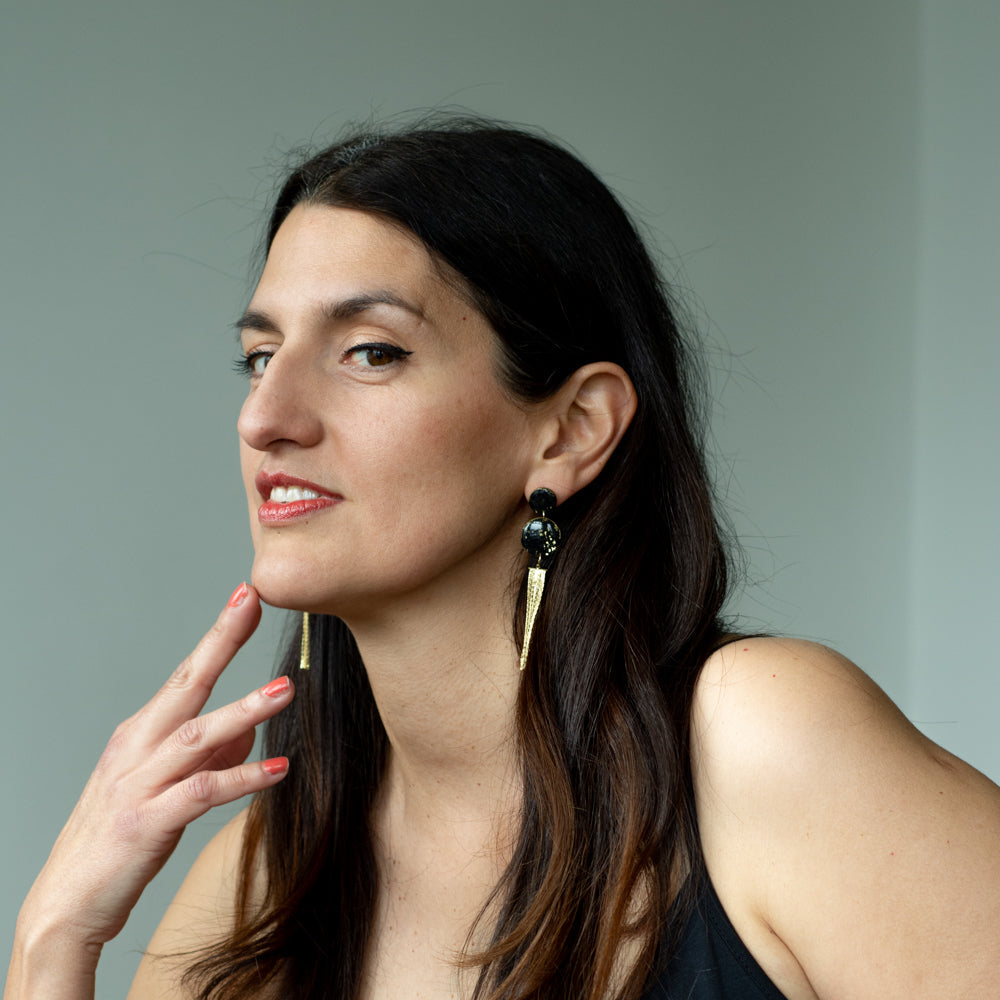 Model wearing handmade black and gold tassel drop earrings with speckled polymer clay discs and gold-plated chain fringe, elegant statement earrings shown in profile.