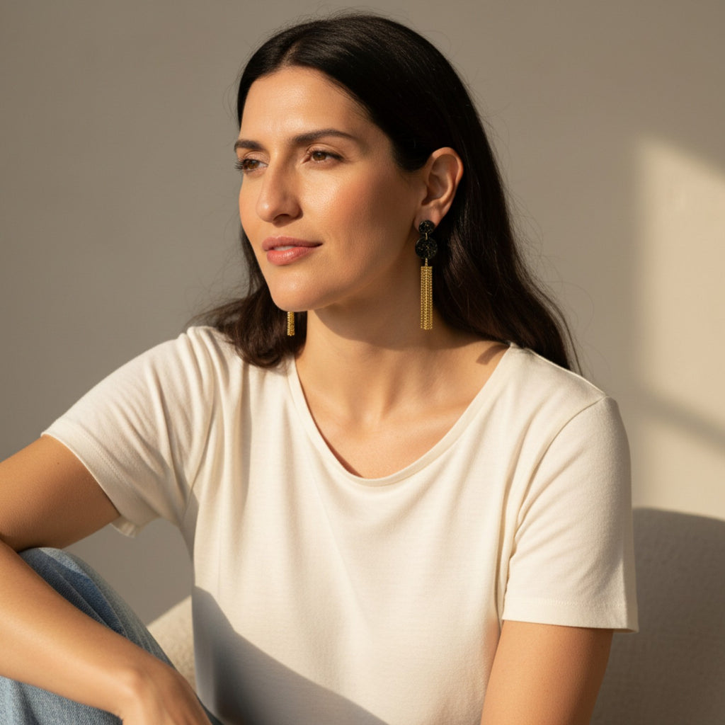 Woman wearing a white t-shirt and black and gold earrings with tassels against a neutral background