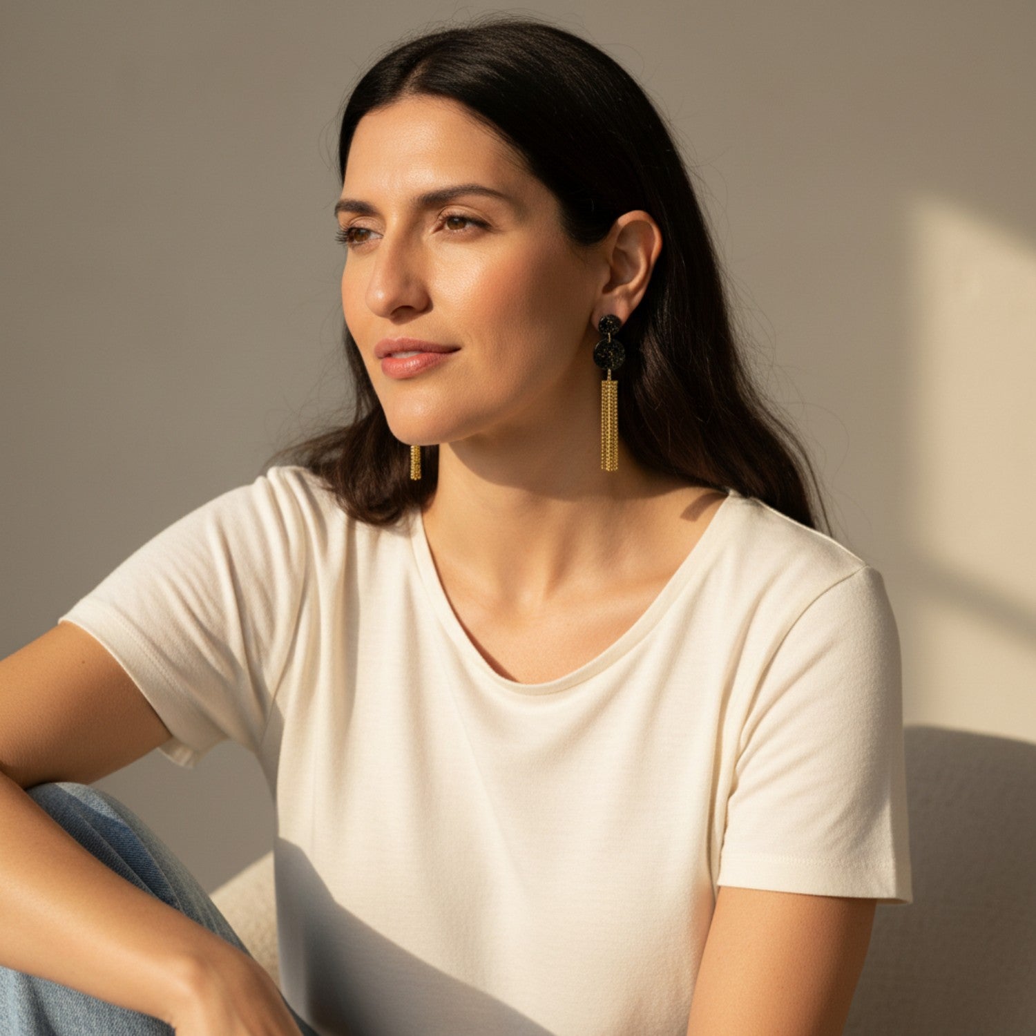 Woman wearing a white t-shirt and black and gold earrings with tassels against a neutral background