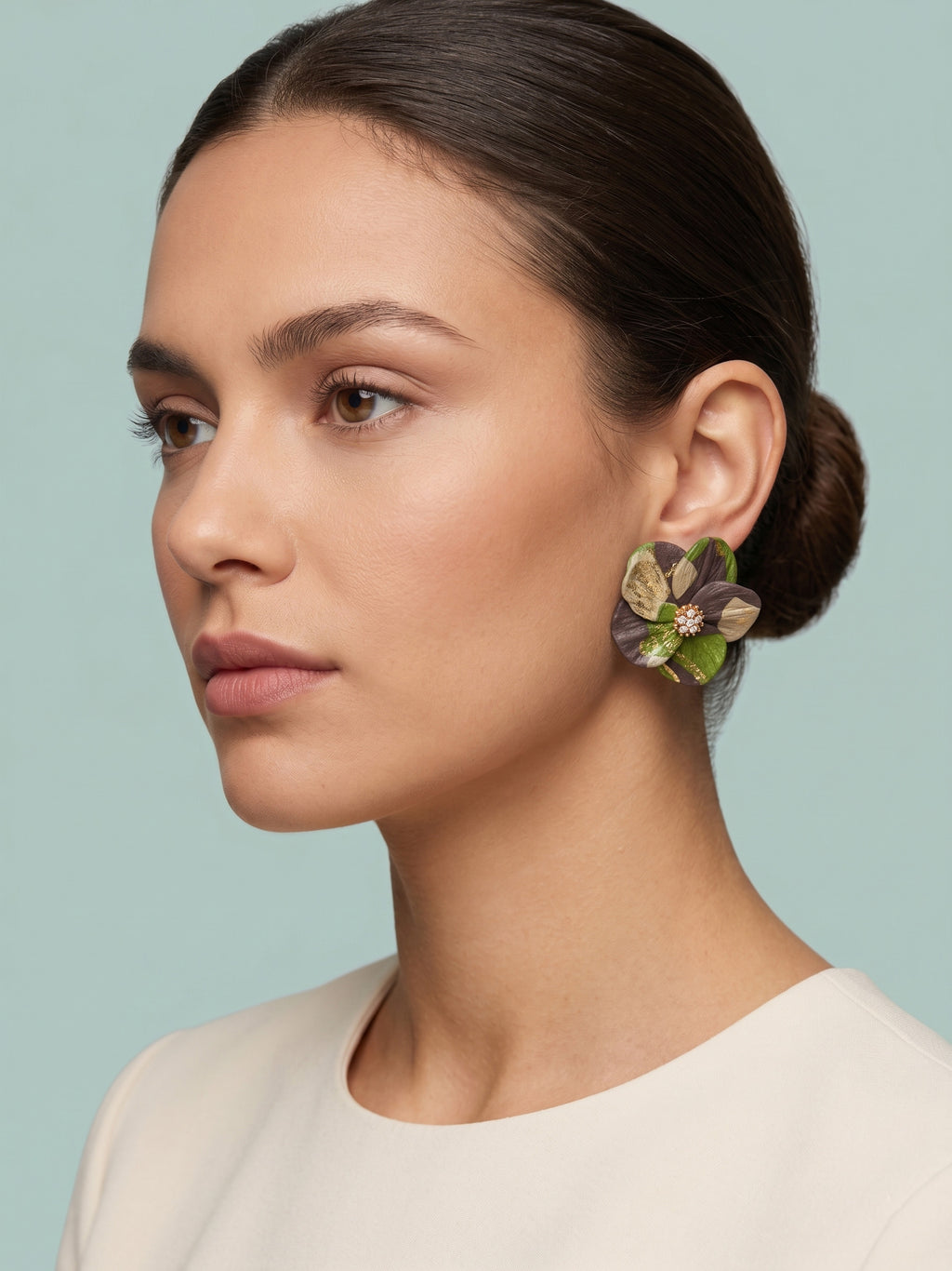A woman in profile wearing a large, sculptural flower-shaped earring with green, purple, and gold petals, set against a solid light blue background.