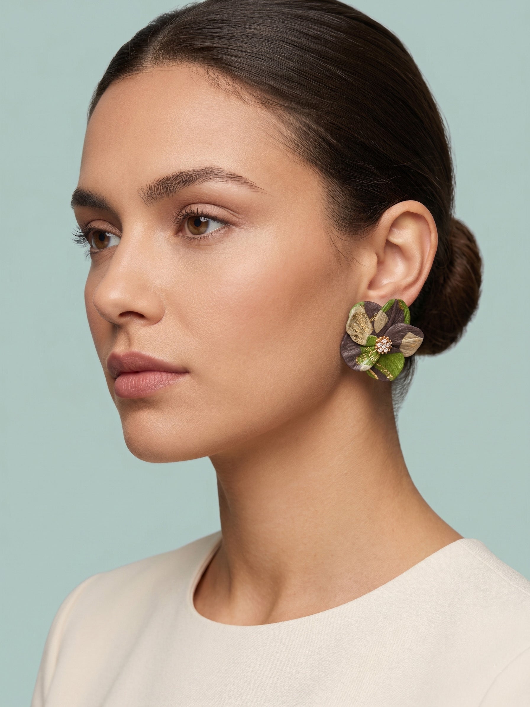 A woman in profile wearing a large, sculptural flower-shaped earring with green, purple, and gold petals, set against a solid light blue background.