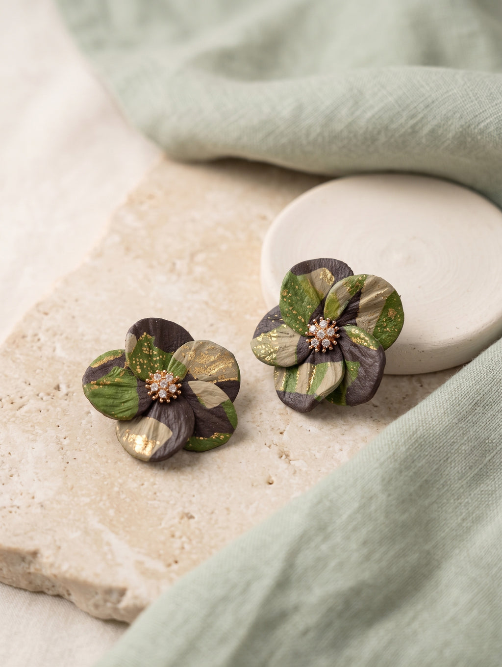 A pair of handmade flower-shaped earrings with green, gold, and dark brown petals, displayed on a stone slab with a sage green cloth and a white ceramic dish in the background.