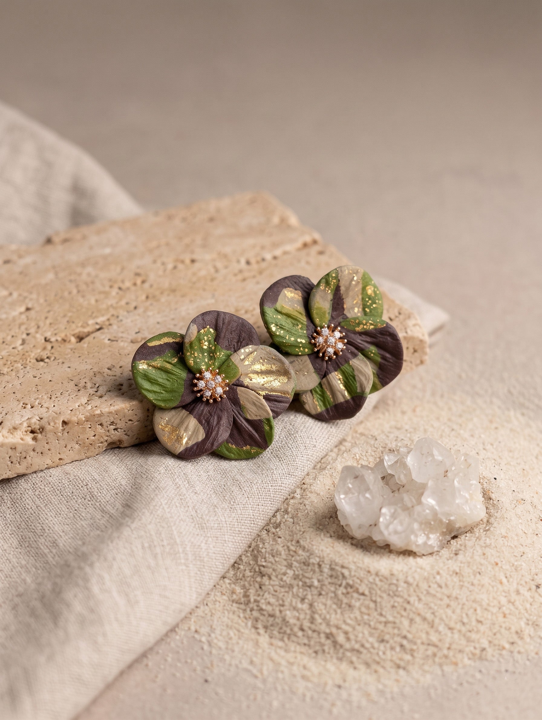 Two handmade flower-shaped earrings with dark purple and olive green petals, metallic gold accents, and small pearl-like centers, displayed on a textured stone slab over a neutral fabric and sand background.