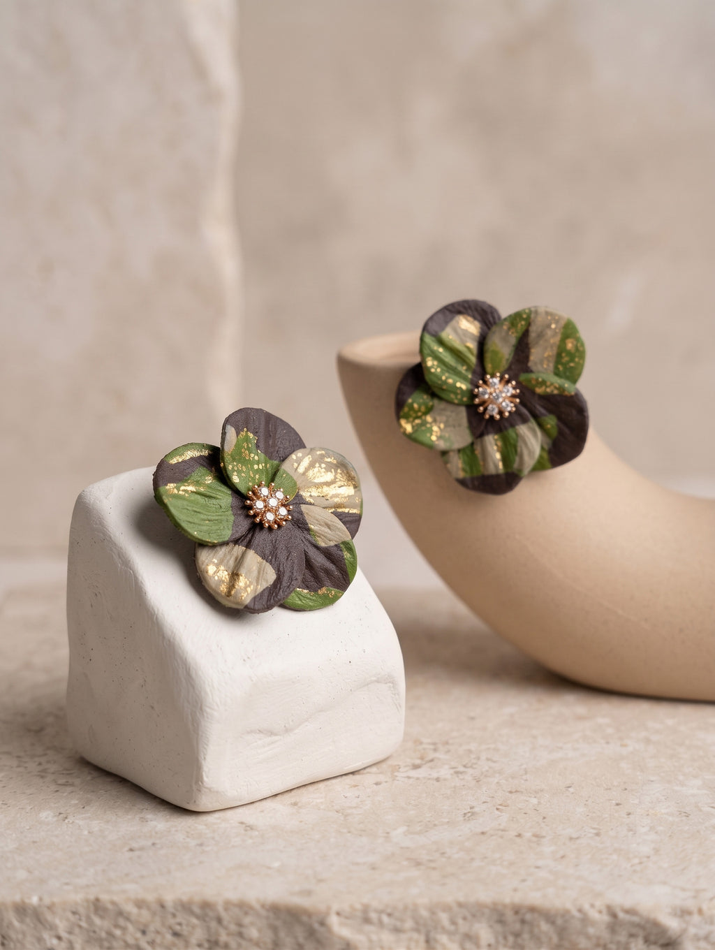 Two handmade flower-shaped earrings with dark purple, green, and metallic gold petals, displayed on a textured beige surface with a ceramic stand.