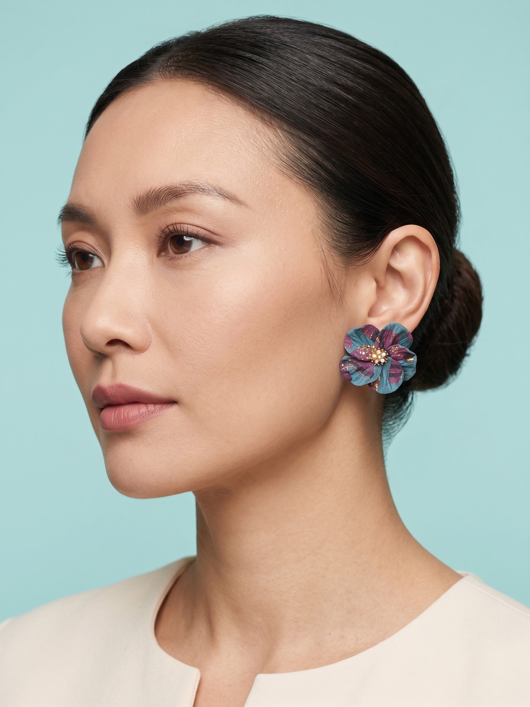 A woman with dark hair in a low bun wears a colorful flower-shaped earring with blue, purple, and pink petals and a gold center, set against a light blue background.