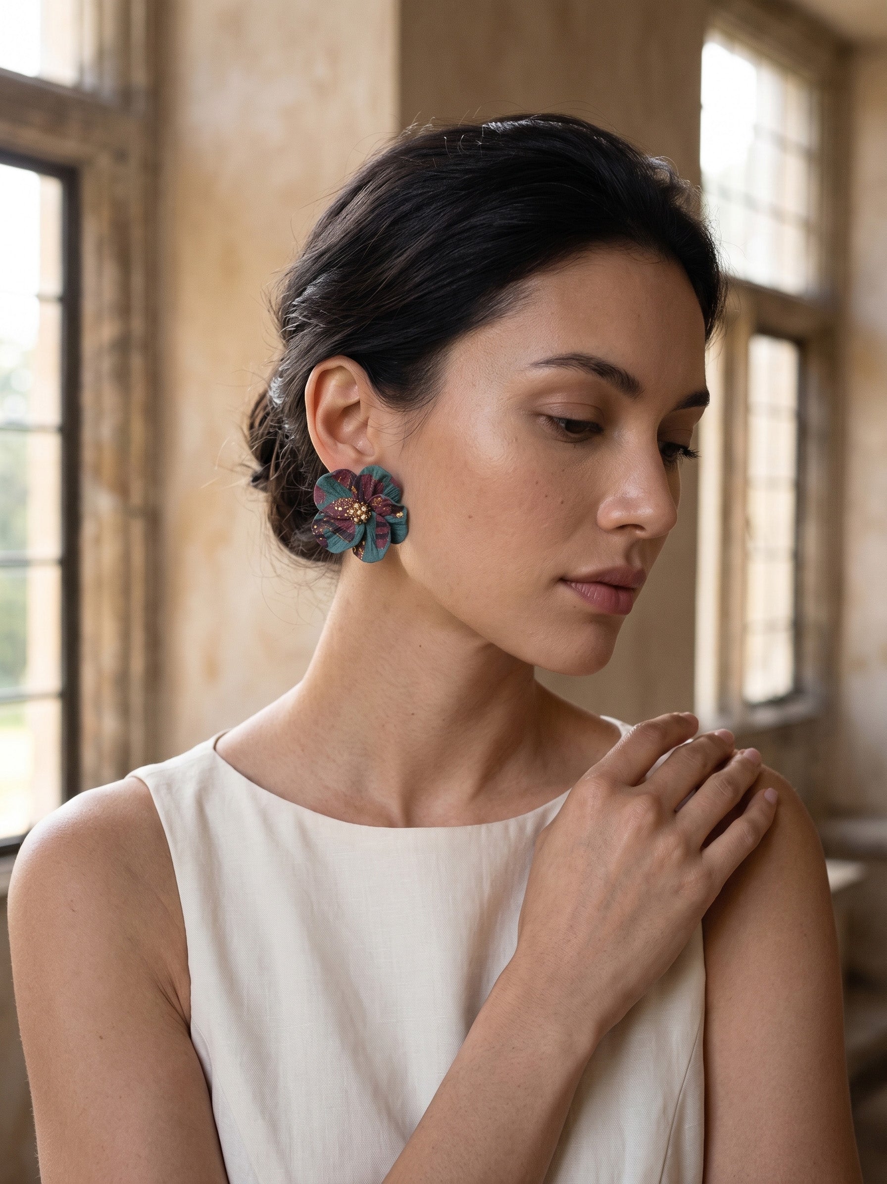 A woman wearing a pair of colorful floral stud earrings with green, pink, and blue tones, styled with a cream-colored sleeveless top in a warm, sunlit interior setting.