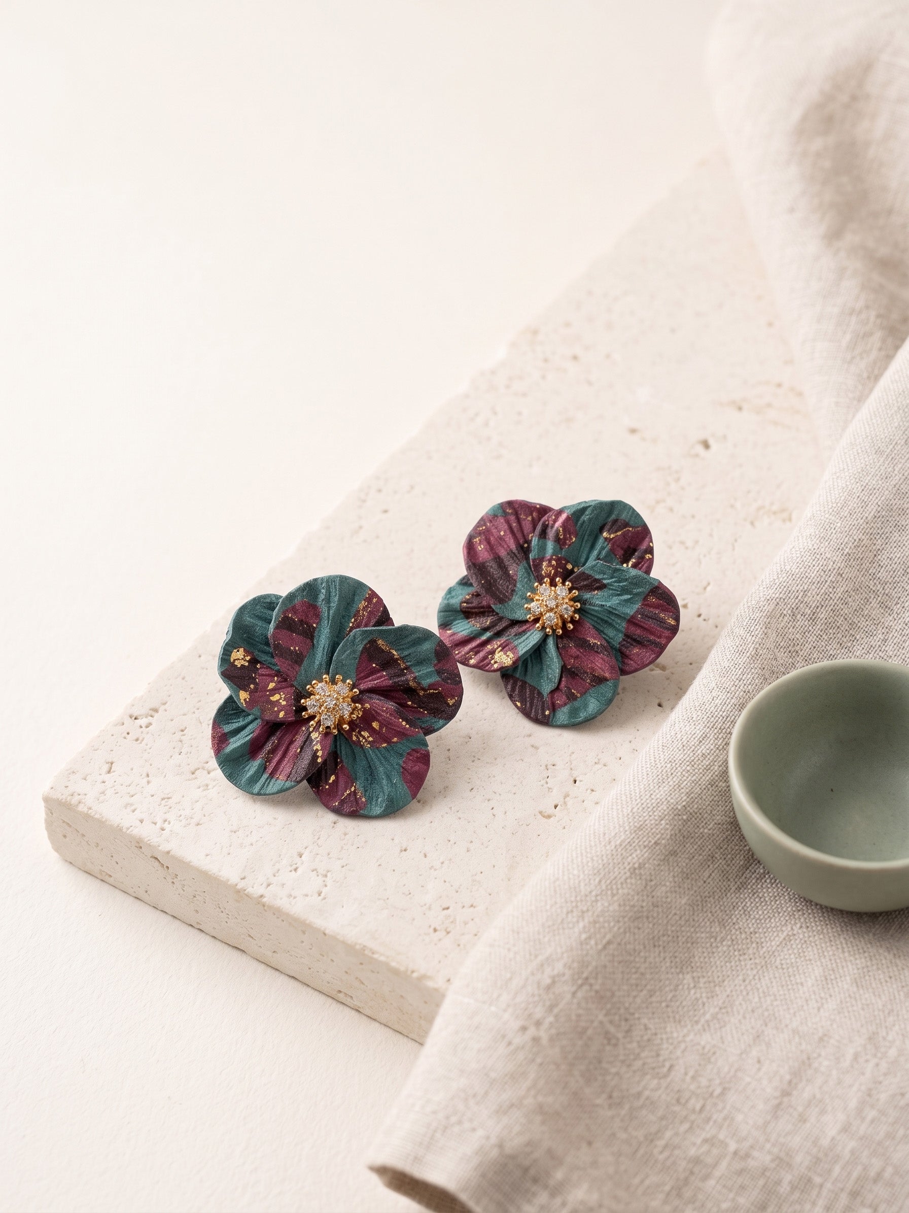 A pair of handmade flower-shaped earrings with layered teal and burgundy petals and a gold center, displayed on a textured stone slab next to a small ceramic cup on a neutral fabric background.