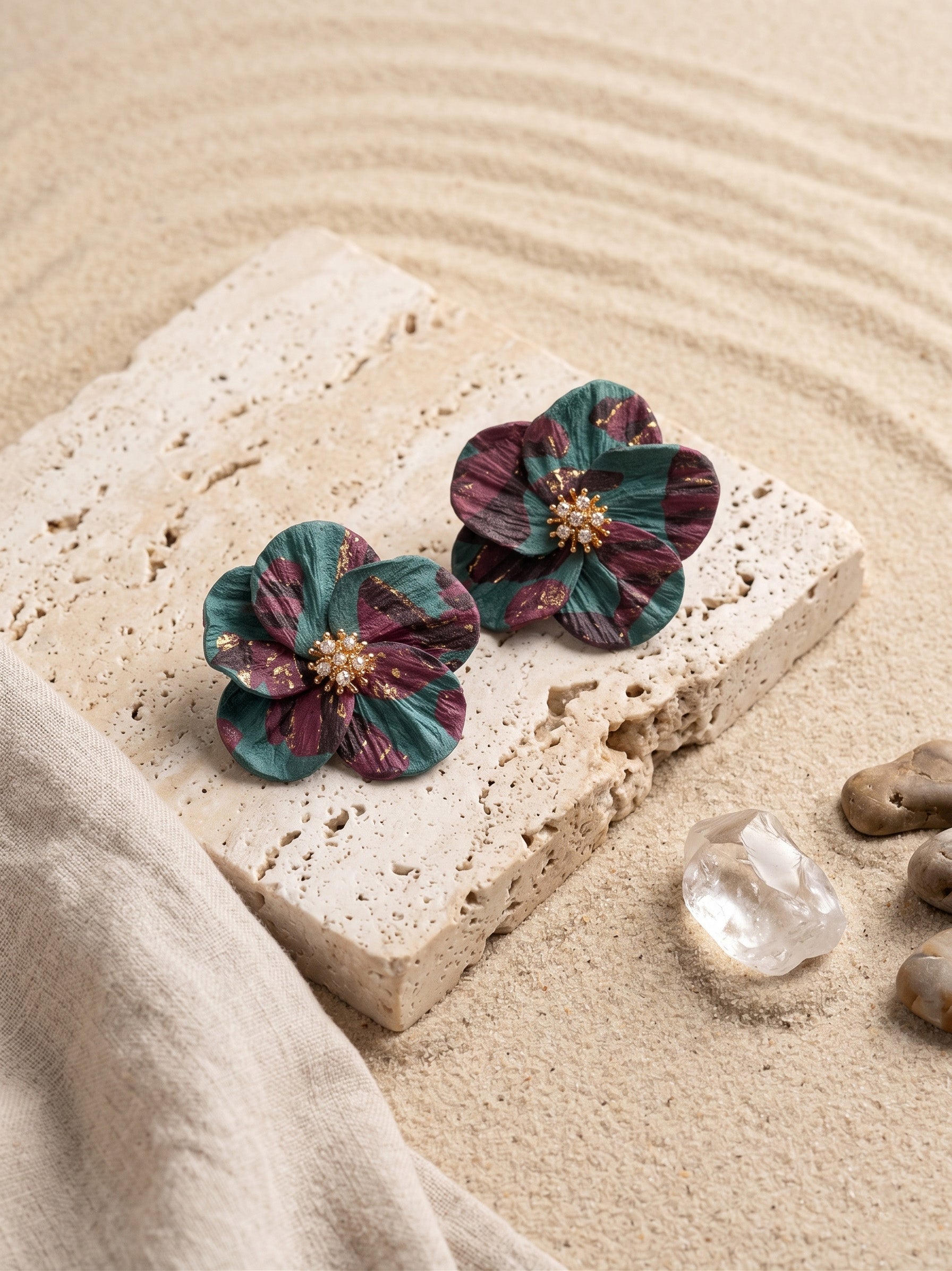 A pair of handmade flower-shaped earrings with marbled teal and burgundy petals and a gold-toned center, displayed on a textured stone slab over a sandy surface with small stones and fabric.