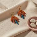 Orange and blue tiered earrings on a beige fabric background with a small pink dish containing stones.
