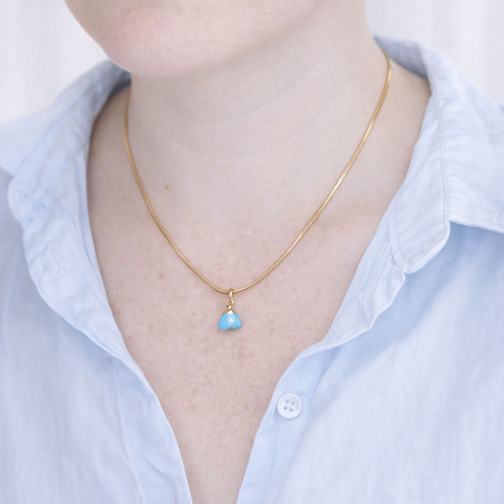 Gold necklace with a turquoise pendant worn by a person in a light blue shirt.