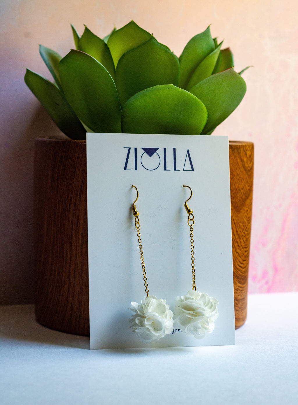 Pair of white pompom earrings on a beige card with a pink bowl in the background