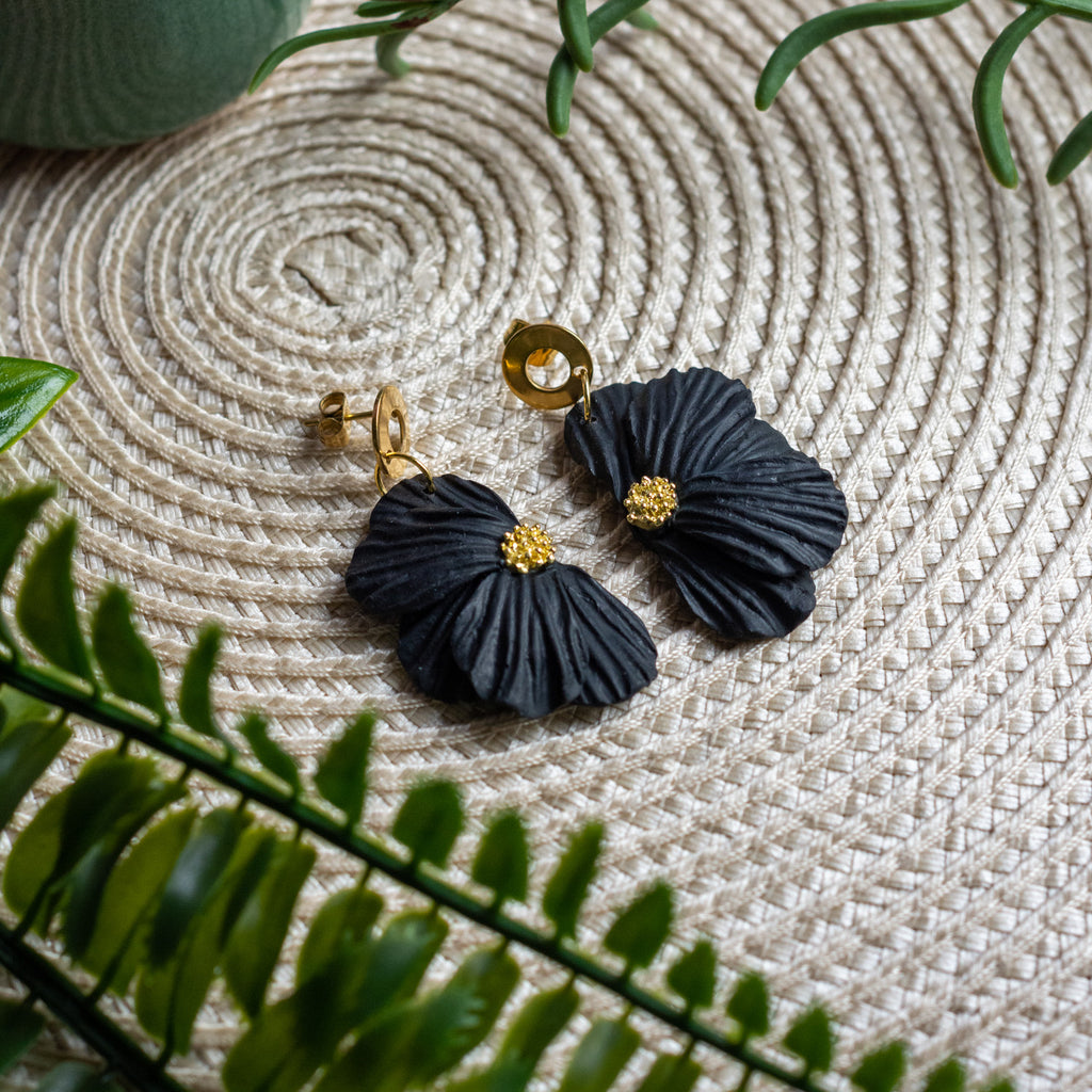 Black flower earrings with gold pistils on a textured surface with green leaves.