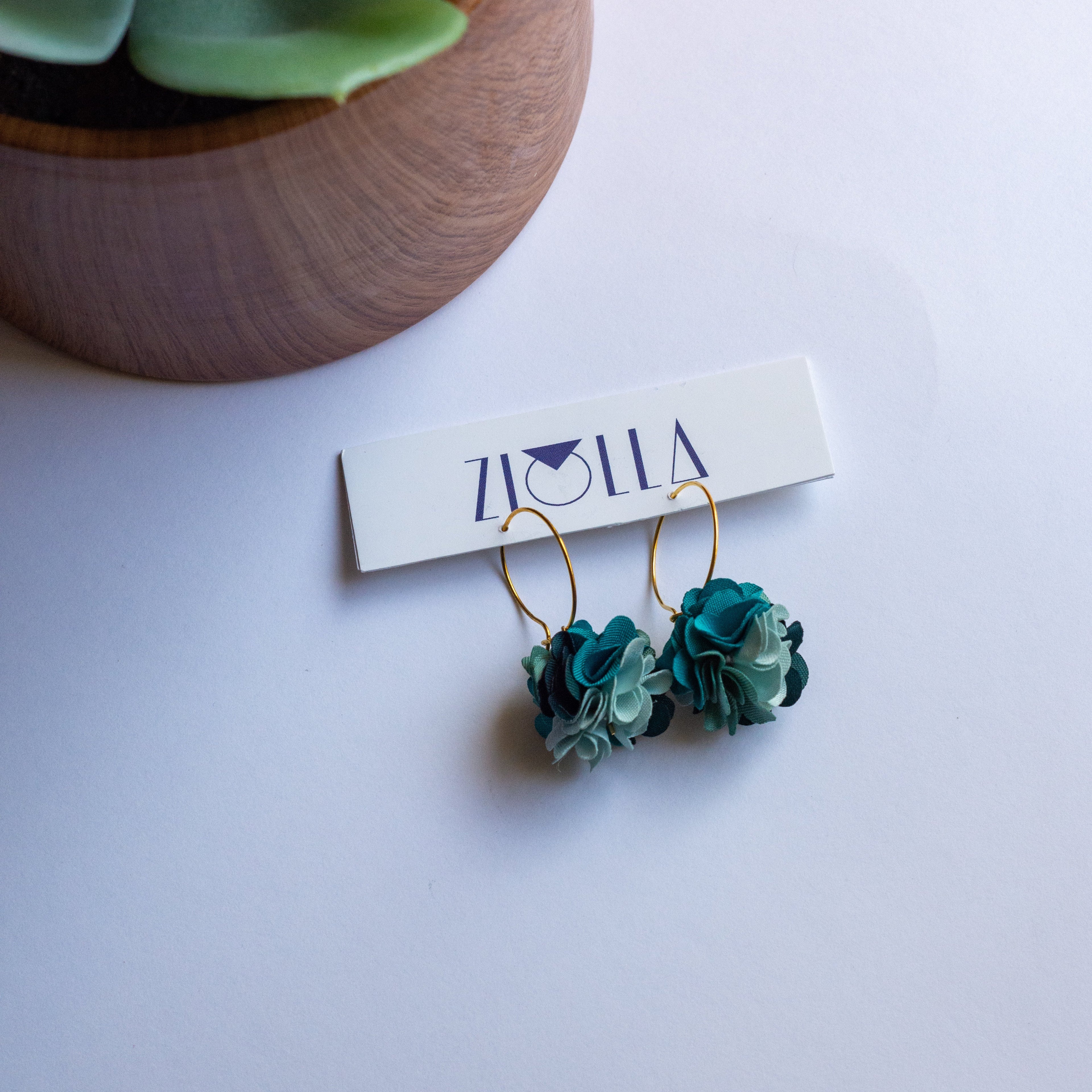 Teal flower-shaped earrings on a white surface with a Zoola card in the background.